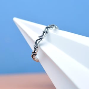 May include: A silver ring featuring a detailed, repeating design of curved lines and tiny dots. The ring is displayed on a white geometric form, set against a backdrop of blue and peach, offering a striking contrast.