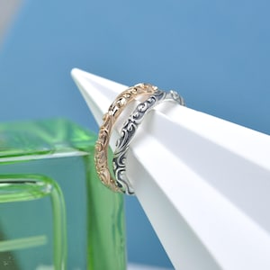 May include: Two decorative rings, one gold and one silver, are presented on a white geometric shape against a blue backdrop. The rings have a repeating, swirling design. A green glass object is visible in the lower left corner.