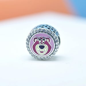 Puede incluir: Un charm de plata con la cara de un oso de dibujos animados en tonos rosa, morado y verde. La cara del oso está centrada en un círculo rosa, rodeado de cristales transparentes. El charm tiene un barril azul oscuro estampado.