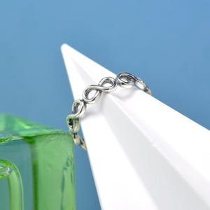 May include: A silver ring with a repeating infinity symbol pattern. The ring is displayed against a white and blue backdrop, with a green object in the foreground. The ring's design is detailed and intricate, showcasing a delicate aesthetic.