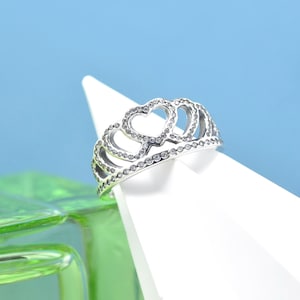 May include: A silver-colored ring in a tiara design, featuring heart-shaped cutouts and small, clear gemstones. The ring is displayed on a white surface against a blue background, showcasing its intricate details.