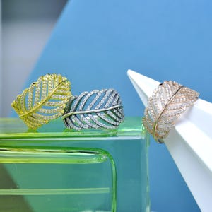 Op de afbeelding: Drie bladvormige ringen, elk bezet met kleine, heldere edelstenen. Eén ring is goudkleurig, een andere is zilverkleurig en de derde is roségoud. De ringen worden getoond op een groen oppervlak tegen een blauwe achtergrond.