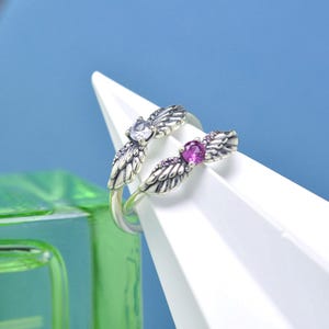 Puede incluir: Un anillo de plata con un dise&ntilde;o abierto, con dos acentos en forma de ala. Un ala tiene una gema transparente, la otra una gema rosa. El anillo se muestra sobre una superficie triangular blanca sobre un fondo azul.