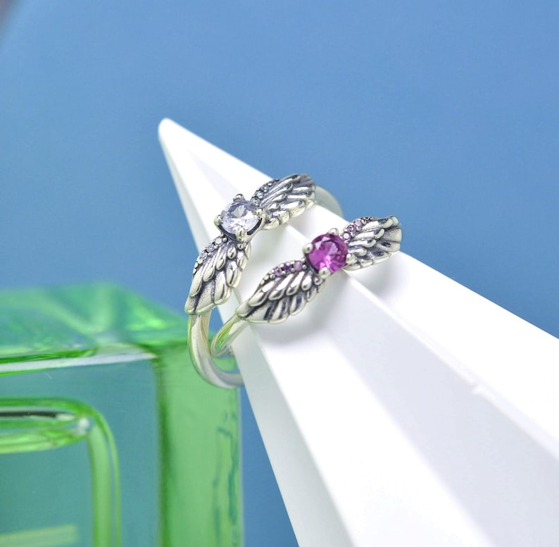 Puede incluir: Un anillo de plata con un dise&ntilde;o abierto, con dos acentos en forma de ala. Un ala tiene una gema transparente, la otra una gema rosa. El anillo se muestra sobre una superficie triangular blanca sobre un fondo azul.