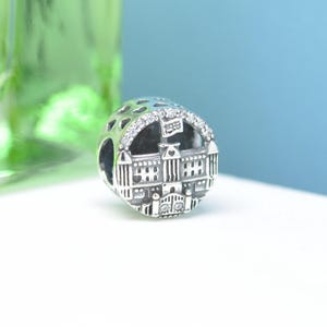 May include: A silver charm with a detailed building design, featuring a flag and sparkling accents. The charm has a round shape with cut-out heart shapes. The building has a detailed facade.