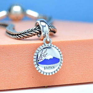 May include: A silver charm with a round pendant featuring a blue mountain, cherry blossoms, and the word "JAPAN". The charm is attached to a silver bracelet. The pendant has a beaded border.