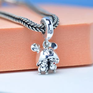 Puede incluir: Un charm de plata con un tema de Mickey Mouse. El charm presenta dos figuras con narices rojas y un sombrero de Mickey Mouse. El charm está unido a una cadena de plata. El fondo es de color naranja claro.