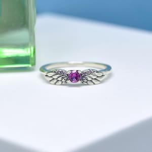 Puede incluir: Un anillo de plata con una piedra preciosa central magenta y redonda. El anillo presenta dise&ntilde;os en forma de alas a cada lado de la piedra preciosa, con piedras magenta m&aacute;s peque&ntilde;as que acent&uacute;an las alas. El anillo est&aacute; sobre una superficie blanca.