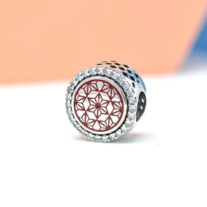 May include: A silver charm with a red and white geometric design. The charm is round with a ring of clear stones around the edge. The charm has a detailed pattern and is likely a jewelry component.