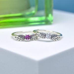 Puede incluir: Dos anillos de plata con dise&ntilde;os de alas. Uno tiene una gema rosa, el otro una transparente. Las alas presentan detalles de plumas. Los anillos est&aacute;n sobre un fondo blanco, con un fondo verde borroso.