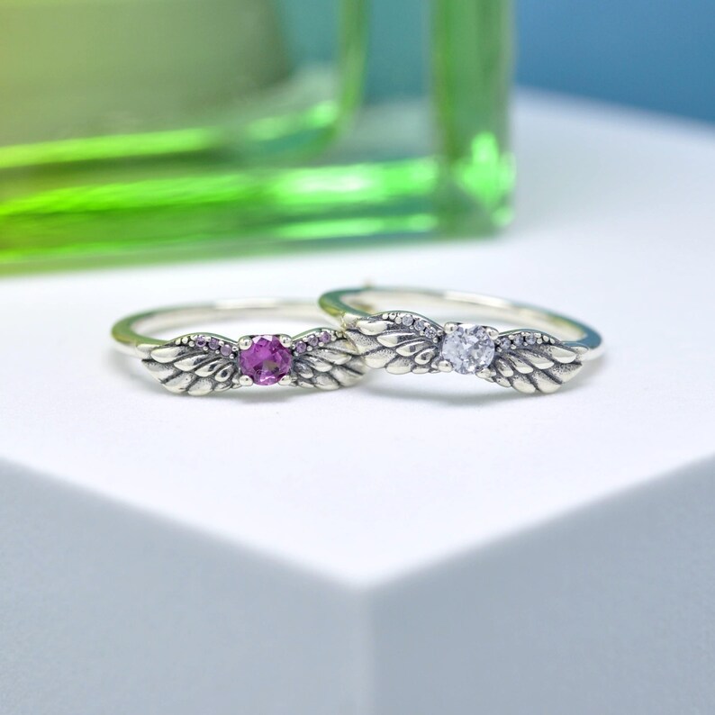 Puede incluir: Dos anillos de plata con dise&ntilde;os de alas. Uno tiene una gema rosa, el otro una transparente. Las alas presentan detalles de plumas. Los anillos est&aacute;n sobre un fondo blanco, con un fondo verde borroso.