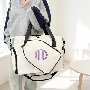 K&ouml;nnte beinhalten: Cremefarbene Reisetasche mit schwarzen Griffen und Zierleisten. Die Tasche hat ein gro&szlig;es, gesticktes lila Monogramm in einem schwarz umrandeten Design. Sie hat einen Schultergurt und eine Seitentasche mit Rei&szlig;verschluss.
