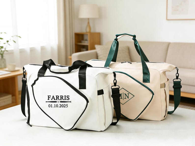 K&ouml;nnte beinhalten: Zwei Reisetaschen. Eine ist wei&szlig; mit schwarzen Verzierungen und dem Namen "FARRIS" und dem Datum "01.10.2025" aufgedruckt. Die andere ist beige mit gr&uuml;nen Verzierungen und den Initialen "N" in einem Diamanten. Beide haben Schultergurte.