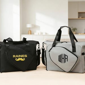K&ouml;nnte beinhalten: Zwei Reisetaschen, eine schwarze und eine graue, stehen nebeneinander. Die schwarze Tasche hat "RAINES" und einen gelben Schnurrbart. Die graue Tasche zeigt "WXR" in Schwarz. Beide Taschen haben schwarze Griffe und Schultergurte.