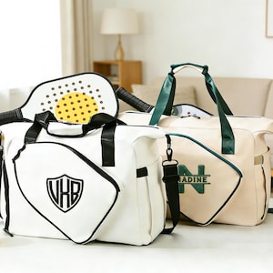 Op de afbeelding: Twee sporttassen, één wit met zwarte randen en een schildlogo, de andere beige met groene accenten en de naam "NADINE". Een pickleball racket en bal zijn ook aanwezig.