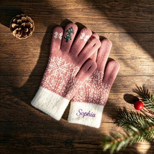 Può includere: Un paio di guanti in maglia rosa polvere con un motivo a fiocchi di neve bianco sul dorso della mano, un piccolo dettaglio a forma di albero verde sul dito medio e un polsino bianco. Il nome Sophia è ricamato in viola sul polsino destro.