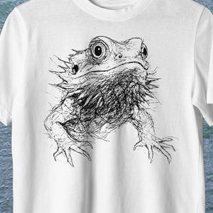 Peut inclure: T-shirt blanc avec une illustration en noir et blanc d'un dragon barbu. Le t-shirt est suspendu à un cintre en bois. Le texte "WHITE COLOR" est visible en haut à droite.