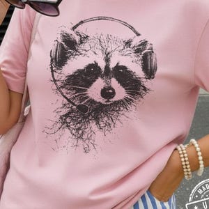 May include: Pink t-shirt with a graphic of a raccoon wearing a headset. The raccoon is depicted in black and white, with detailed fur and facial features. The shirt has short sleeves and a crew neck. The text "BLOSSOM COLOR" is printed on the bottom.