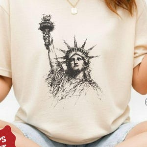 May include: Ivory-colored t-shirt featuring a detailed black and white sketch of the Statue of Liberty. The shirt has short sleeves and a crew neck. The text "IVORY COLOR" is printed at the top left.