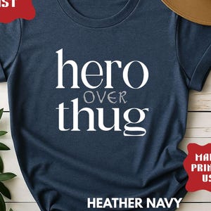 Peut inclure: T-shirt bleu marine avec l'inscription blanche "hero over thug". Le t-shirt a des manches retroussées et est présenté sur une surface en bois blanc. Un chapeau marron est visible en arrière-plan.