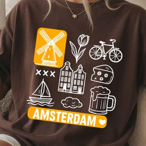 Espresso colored t-shirt with white graphic icons of a windmill, tulips, bicycle, houses, cheese, beer, and a sailboat. The word "AMSTERDAM" is in orange.