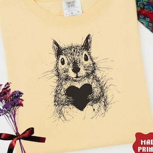 Peut inclure: Sweat-shirt de couleur banane avec un dessin de l'écureuil noir tenant un cœur. Le texte "BANANA COLOR" est dans le coin supérieur gauche, et "MADE / PRINTED USA" est dans le coin inférieur droit. Un bouquet floral et un foulard à motifs sont également dans l'image.