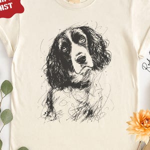May include: An ivory-colored short-sleeve t-shirt featuring a black ink-style sketch of a spaniel dog on the front. The graphic depicts the dog's head and chest with expressive, loose lines. The shirt has a crew neckline and a standard fit.
