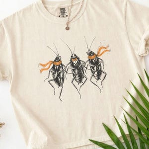 Puede incluir: Una camiseta de manga corta color marfil con una ilustración gráfica de tres cucarachas de pie. Dos de los insectos llevan bufandas naranjas y el del centro lleva una pajarita naranja. El texto IVORY está impreso en la esquina superior izquierda.