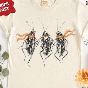 May include: An ivory-colored t-shirt featuring a black ink illustration of three cockroaches standing in a row. The outer two insects wear orange scarves, while the center one wears a bow tie. The shirt includes text reading IVORY COLOR.