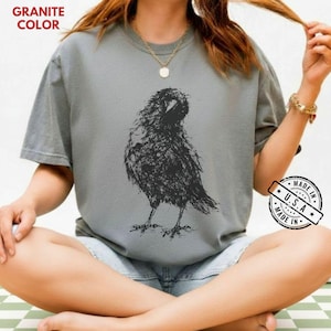 May include: A granite-colored short-sleeve t-shirt featuring a black, sketch-style graphic of a crow in the center. The shirt has a crew neckline and is labeled with the text GRANITE COLOR in the top left corner and a MADE IN U.S.A. circular badge.