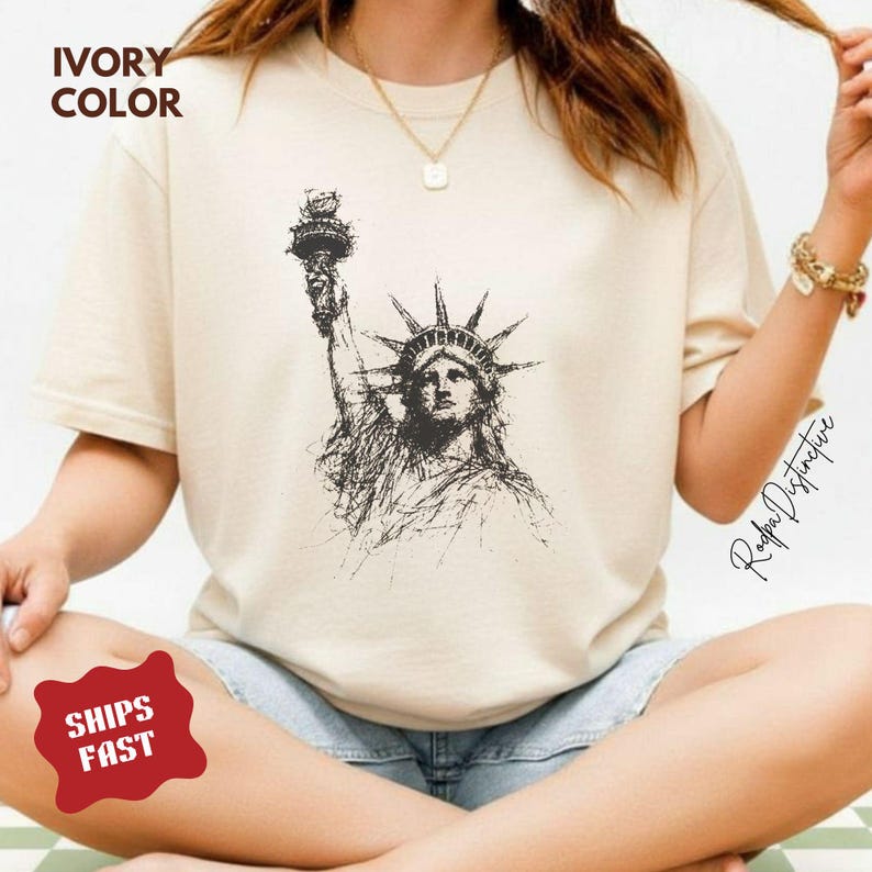 May include: Ivory-colored t-shirt featuring a detailed black and white sketch of the Statue of Liberty. The shirt has short sleeves and a crew neck. The text "IVORY COLOR" is printed at the top left.