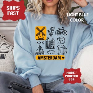 Pu&ograve; includere: Felpa azzurra con grafica di Amsterdam nera e gialla. Il design include un mulino a vento, un tulipano, una bicicletta, case, formaggio, un boccale di birra, una barca a vela e la scritta "AMSTERDAM".
