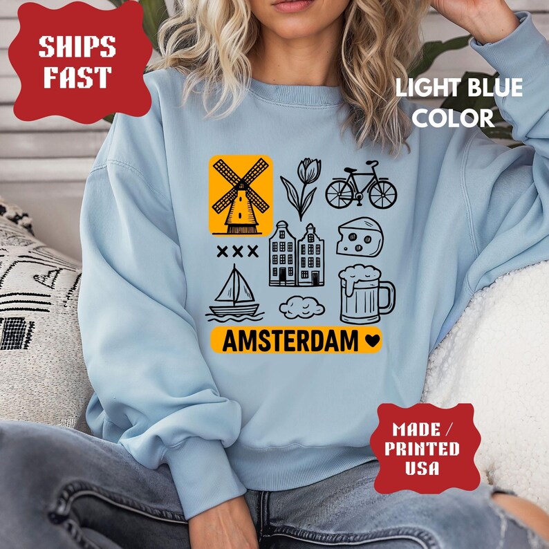 Pu&ograve; includere: Felpa azzurra con grafica di Amsterdam nera e gialla. Il design include un mulino a vento, un tulipano, una bicicletta, case, formaggio, un boccale di birra, una barca a vela e la scritta "AMSTERDAM".