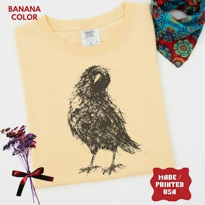 May include: A banana-colored t-shirt featuring a black, sketch-style graphic of a crow. The shirt has a crew neckline and a white interior label. The text BANANA COLOR is in the top left, and MADE / PRINTED USA is in the bottom right.