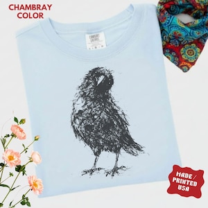 May include: A light blue t-shirt in chambray color featuring a black ink-style sketch of a bird. The shirt has a crew neckline and a white interior label. The graphic depicts a bird with textured, scratchy lines. Text on the image reads CHAMBRAY COLOR and MADE / PRINTED USA.