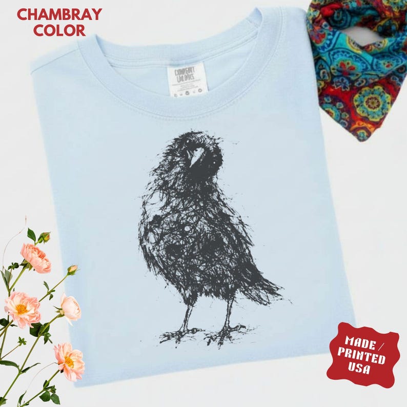 May include: A light blue t-shirt in chambray color featuring a black ink-style sketch of a bird. The shirt has a crew neckline and a white interior label. The graphic depicts a bird with textured, scratchy lines. Text on the image reads CHAMBRAY COLOR and MADE / PRINTED USA.