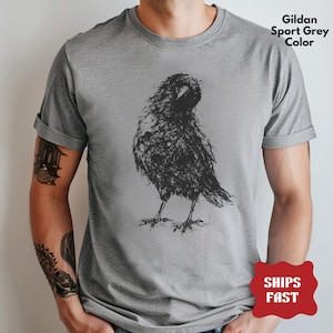 May include: A short-sleeved crew neck t-shirt in Gildan Sport Grey color. The front features a black graphic illustration of a bird, rendered in a sketchy, scribbled line art style. The bird is depicted standing in profile with its head turned.