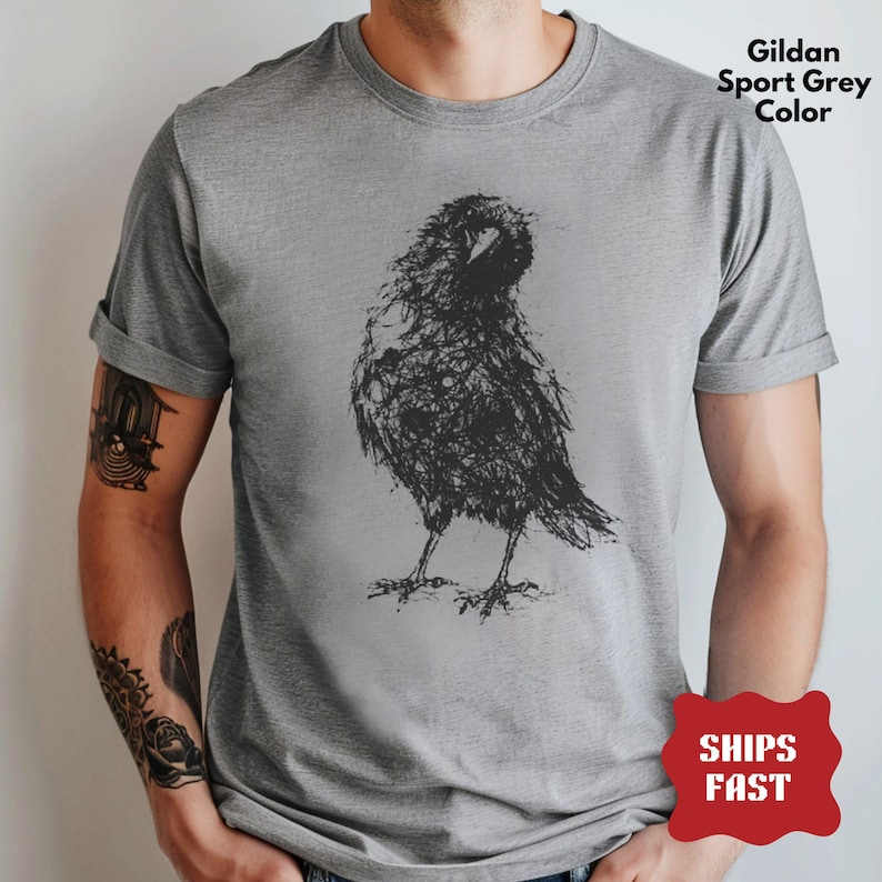 May include: A short-sleeved crew neck t-shirt in Gildan Sport Grey color. The front features a black graphic illustration of a bird, rendered in a sketchy, scribbled line art style. The bird is depicted standing in profile with its head turned.