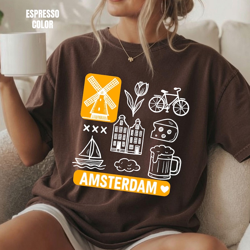 Espresso colored t-shirt with white graphic icons of a windmill, tulips, bicycle, houses, cheese, beer, and a sailboat. The word "AMSTERDAM" is in orange.