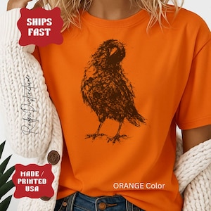May include: A bright orange short-sleeved t-shirt featuring a dark brown, sketch-style graphic of a bird resembling a crow or raven in the center. The shirt is worn under a white, chunky knit cardigan with visible brown buttons on the front.