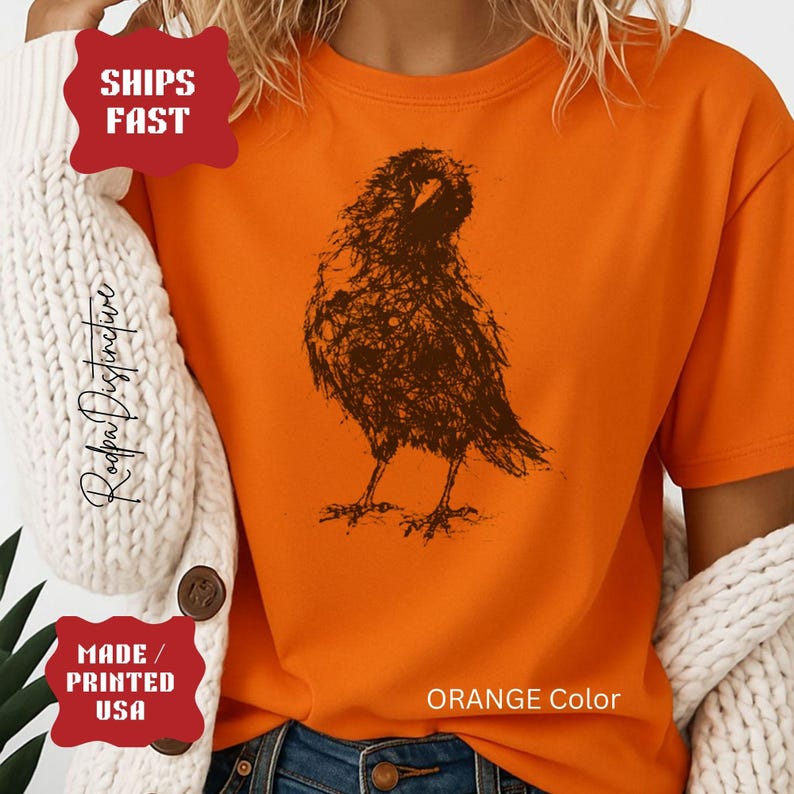 May include: A bright orange short-sleeved t-shirt featuring a dark brown, sketch-style graphic of a bird resembling a crow or raven in the center. The shirt is worn under a white, chunky knit cardigan with visible brown buttons on the front.