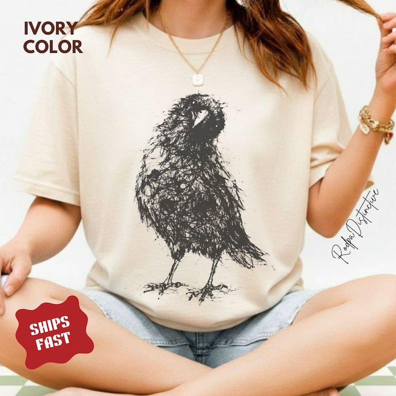 May include: An ivory-colored short-sleeve t-shirt featuring a black graphic illustration of a bird created with a scribbled, sketch-like line art style. The shirt has a crew neckline and a relaxed fit, showcasing a detailed, textured bird design.