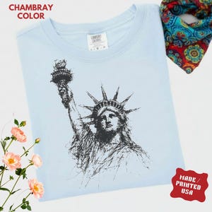 May include: Light blue t-shirt with a black sketch of the Statue of Liberty. The shirt has the text "CHAMBRAY COLOR" and "MADE / PRINTED USA". A colorful paisley scarf is in the background.