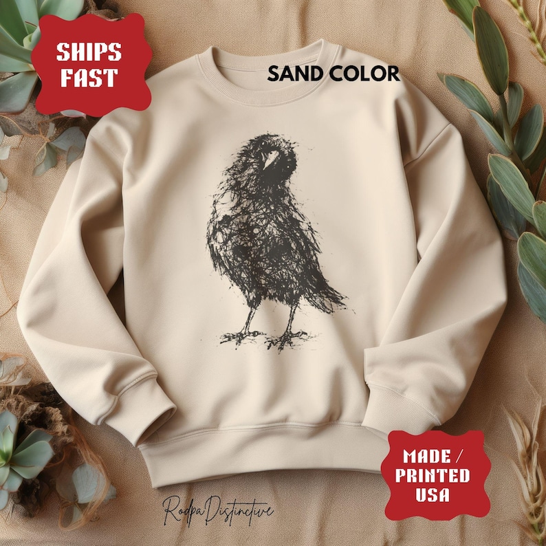 May include: A sand-colored crewneck sweatshirt featuring a black ink-style sketch of a raven on the front. The garment includes text that reads SHIPS FAST, SAND COLOR, and MADE / PRINTED USA, along with the brand name Rodpa Distinctive.
