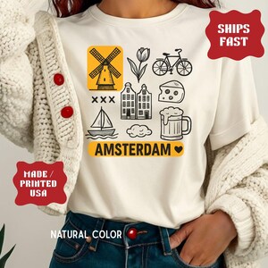 Pu&ograve; includere: T-shirt color crema con grafica a tema Amsterdam in bianco e giallo. Il design include un mulino a vento, un tulipano, una bicicletta, case, formaggio, birra e una barca a vela. La parola "AMSTERDAM" &egrave; stampata in giallo.