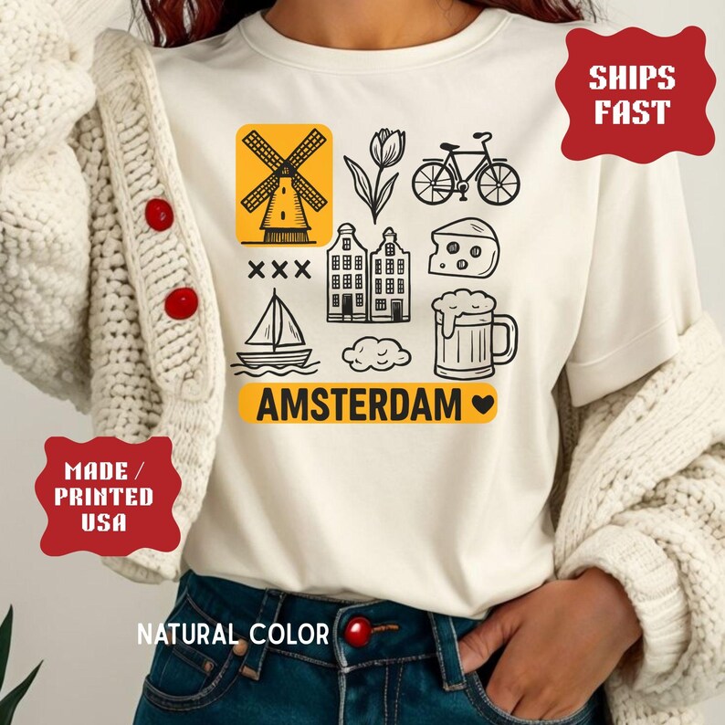 Pu&ograve; includere: T-shirt color crema con grafica a tema Amsterdam in bianco e giallo. Il design include un mulino a vento, un tulipano, una bicicletta, case, formaggio, birra e una barca a vela. La parola "AMSTERDAM" &egrave; stampata in giallo.
