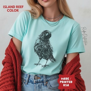 May include: An island reef colored t-shirt featuring a dark charcoal graphic of a raven created with a scribbled, sketch-like line art style. The shirt has short sleeves and a crew neckline. Text on the image reads ISLAND REEF COLOR and MADE / PRINTED USA.