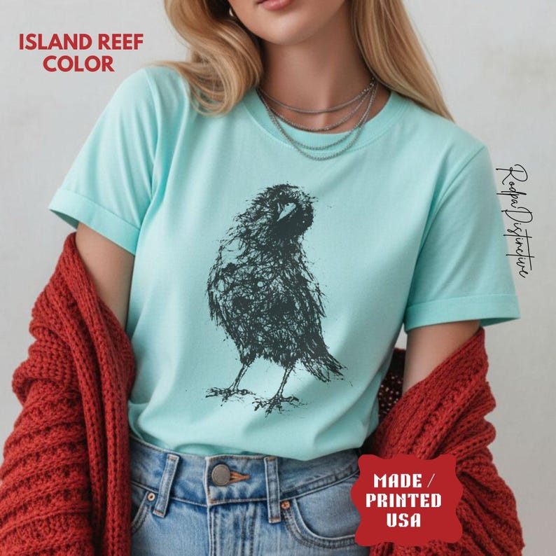 May include: An island reef colored t-shirt featuring a dark charcoal graphic of a raven created with a scribbled, sketch-like line art style. The shirt has short sleeves and a crew neckline. Text on the image reads ISLAND REEF COLOR and MADE / PRINTED USA.