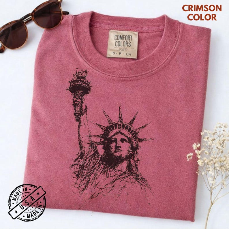 May include: Crimson-colored sweatshirt featuring a sketch of the Statue of Liberty. The shirt has a crew neck and a tag that reads "COMFORT COLORS". The text "MADE IN U.S.A." is printed on a circular sticker. Sunglasses and flowers are also in the image.