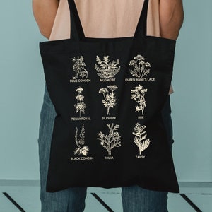 May include: Black tote bag featuring gold botanical illustrations and plant names. The bag displays nine different plant species, including Blue Cohosh, Mugwort, and Queen Anne's Lace. The bag is held by a person.